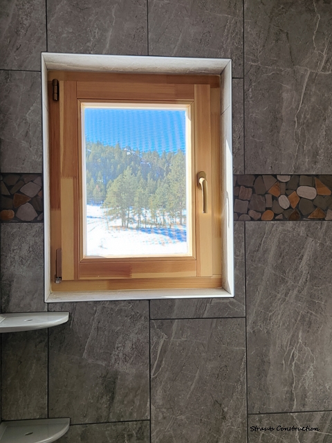 Straub Construction-9 Wood-framed casement window in a gray-tiled bathroom with a river-rock accent stripe, overlooking a snowy Colorado pine forest