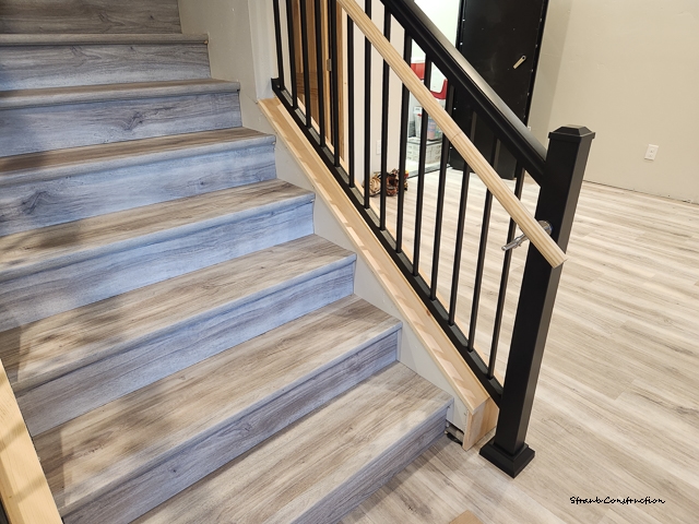 Finished basement staircase with gray plank treads, black metal balusters, and a natural wood handrail