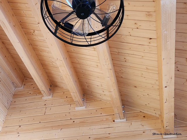 Vaulted tongue-and-groove pine ceiling with exposed beams and a black industrial cage fan in a custom cabin